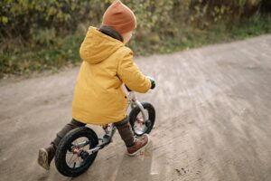 A child wearing a yellow jacket and beanie rides a bike on a dirt path outdoors.